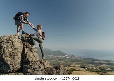 Young Hiker An Holding A Woman Hand Helps To Climb To The Top Of The Mountain. The Concept Of Help And Support. Motivation For Success