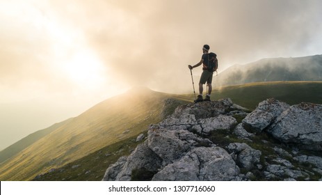 Young hiker man with backpack and walking poles, standing on peak of a mountain looking at sunset in cloudy sky. Green field and rocks. Abruzzo, Italy. - Shutterstock ID 1307716060