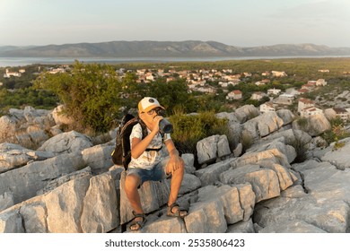 Young hiker wearing cap and drinking water while taking break on rocky terrain during mountain trek. Concept of youth adventure, outdoor exploration, and hiking in nature. High quality photo - Powered by Shutterstock