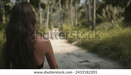 Young hiker woman with long brown hair, back view, walking in jungle forest. Girl hiking alone. Tourist on jungle park trail, looking around. Exotic travel, tourism, holiday, hiking, active lifestyle.