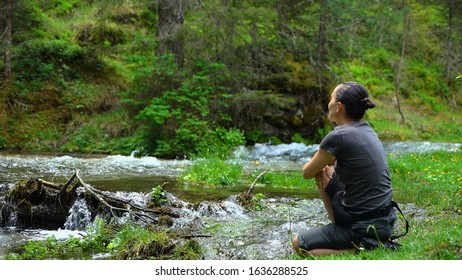 Young hiker woman sitting by a mountain stream, enjoys spending free time at nature