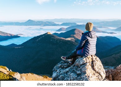 Young Hiker Woman Sitting On The Mountain Summit Cliff And Enjoying Mountains Valley Covered With Clouds View. Successful Summit Climbing Concept Image.