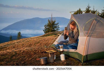 Young hiker woman taking photo with smartphone at dawn in the mountains from her tent next to cooking equipment during hiking. On the backdrop trees and mist at the foot of mountain hills.