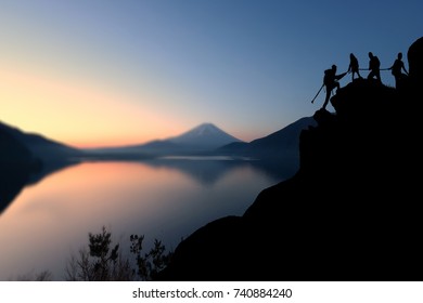 Young Hikers Climbing Up Silhouette Mountain Cliff And One Of Them Giving Helping Hand. Helps, Business Helping And Teamwork Concept. 