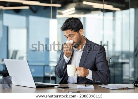 Young Hindu businessman sneezes and has a runny nose while working in the office at the computer, young man has a cold, drinks medicine, feels bad.