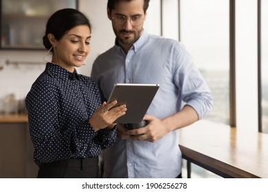 Young hindu female office employee mentor show interested male intern problem solution online using tablet. Two coworkers of different ethnicities discuss work meeting at dining area on lunch break.