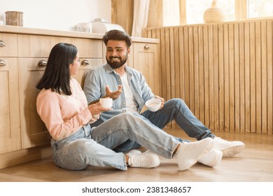 Young Hindu spouses share a light moment while seated on the kitchen floor, engaging in conversation over coffee. Their relaxed posture and smiles show a deep relationship