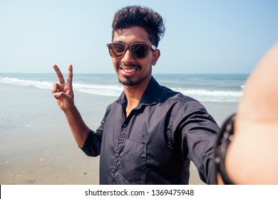 Young Hindu Stylish Man Taking A Picture Self Portrait On The Smartphone Front Camera With Sunglasses Active Beach Vacation On Semmertime Happy Goa India Beach. Sunscreen Spf Protection Concept.