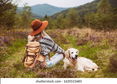 Young Hippie Blond Hair Woman In Wool Sweater With Textile Backpack And Smiling Happy Golden Retriever Dog Sitting On Grass In Summer Mountains Valley. Pets Hiking And Weekend Activities Concept.