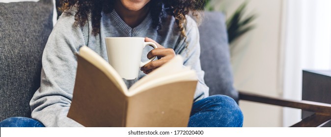 Young hipster african american black woman relaxing reading the paper book work study and looking at page magazine while sitting on couch at home