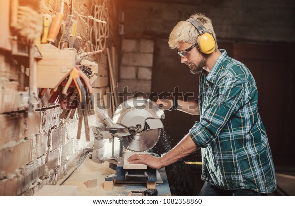 Young hipster bearded man with ear\
protectors by profession carpenter builder saws with a circular saw\
a wooden board on a wooden table in the\
workshop.