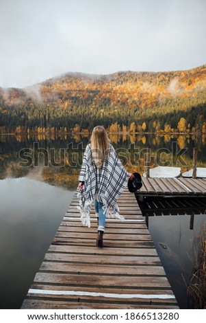 Young hipster blonde woman standing on the pier near the wonderful mountain lake in autumn season at sunrise.
