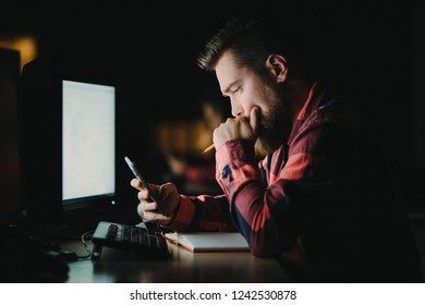 Young Hipster Business Man Holding Cellphone In Front Of The Computer Working Late In Office