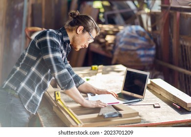 Young Hipster Carpenter Man Working With Computer Laptop In Workshop . Craftsman Wearing Safety Glasses Checking Order Of Clients Or Learning Online