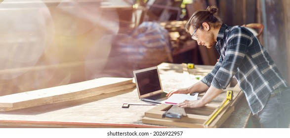 Young Hipster Carpenter Man Working With Computer Laptop In Workshop . Craftsman Wearing Safety Glasses Checking Order Of Clients Or Learning Online