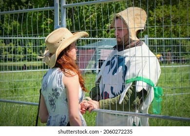 Young Hipster Couple Looking Tenderly At Each Other Over A Wire Fence, Holding Hands.Redhead Girl In Boho Hat Dating With Her Boyfriend During Knightly Tournament Break. Moscow,Russia,May 2019