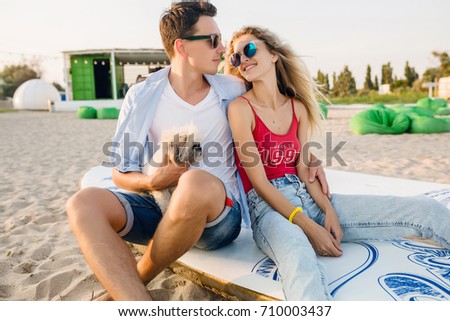 young hipster couple on beach playing with dog Shih Tzu breed, smiling, happy, having fun, summer casual style, romantic mood, family together, sunglasses, surf board, hugging,