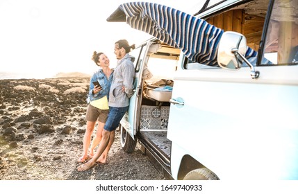 Young hipster couple at relax moment together by oldtimer mini van - Travel lifestyle concept with indie people on minivan adventure trip having fun with mobile smart phone - Warm sunshine filter