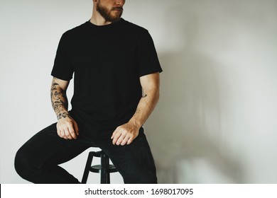 young hipster dressed black t-shirt with empty space for logo, text or design sits on a bar stool against a white wall. mock-up of t-shirt, white wall in the background.
