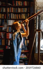 Young Hipster Female Looks Through A Telescope Standing In Vintage Library Room.