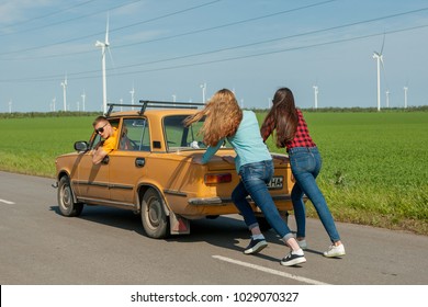 Young hipster friends on road trip on a summers day. Engine break down.Two girls pushing a vintage car while man is emboldening their.Travel, adventure, unforeseenteamwork, funny concept