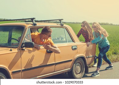 Young Hipster Friends On Road Trip On A Summers Day. Engine Break Down.Two Girls Pushing A Vintage Car While Man Is Emboldening Their.Travel, Adventure, Unforeseenteamwork, Funny Concept