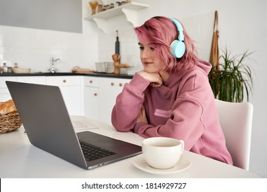 Young Hipster Generation Z Woman Student With Pink Hair Wearing Headphones Looking At Computer Sitting At Kitchen Table, Using Laptop, Learning Online, Watching Webinar, Remote Study In Web At Home.