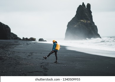 Young hipster girl walks black beach Vík in Iceland, yellow backpack and hat