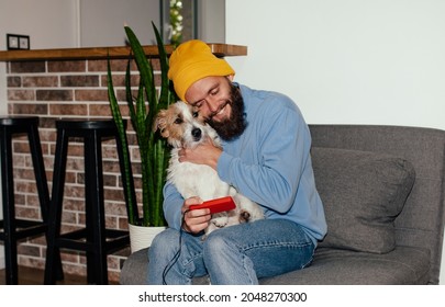 A Young Hipster Guy Sitting On The Couch Playing Computer Games Together With His Dog Jack Russel Terrier. Friendship Between Man And Dog