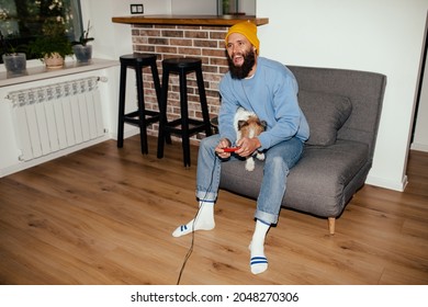 A Young Hipster Guy Sitting On The Couch Playing Computer Games Together With His Dog Jack Russel Terrier. Friendship Between Man And Dog