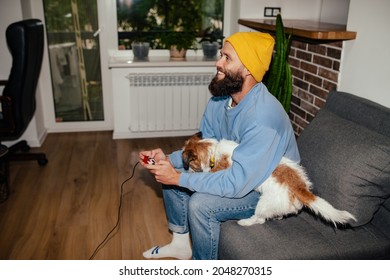 A Young Hipster Guy Sitting On The Couch Playing Computer Games Together With His Dog Jack Russel Terrier. Friendship Between Man And Dog