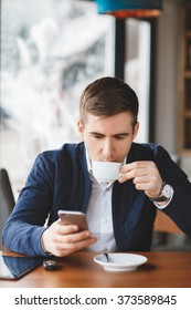 Young Hipster Guy Texting With His Mobile Phone At The Bar And Having A Cappuccino.Young Fashion Man / Hipster Drinking Espresso Coffee In City Cafe During Lunch Time And Working On Tablet Computer