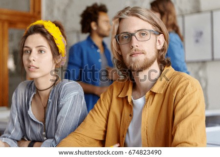 Young hipster male freelancer with beard wearing shirt and eyewear having clever look sitting near his business partner having meeting together discussing main things and plans of their company.