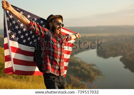 Young hipster man with backpack posing happily holding American flag on background of nature. 