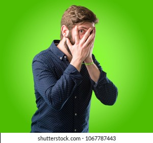 Young hipster man with big beard smiling having shy look peeking through her fingers, covering face with hands looking confusedly broadly over green background