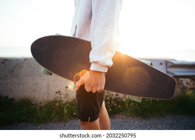 Young hipster man holds cool longboard in hand. Sun sunset light leak in lens. Beautiful travel wanderlust concept. Millennial adventure trip. Skater walk with skateboard 