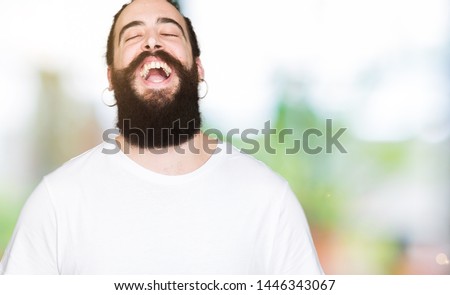 Young hipster man with long hair and beard wearing casual white t-shirt Smiling and laughing hard out loud because funny crazy joke. Happy expression.