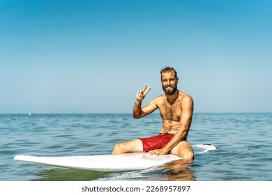 Young hipster man sitting on the surfboard greeting friends with a victory gesture - Guy having fun doing extreme sport - Sporty bearded man training with surfboard on the sea - freedom concept - Powered by Shutterstock