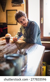 Young Hipster Man Sketching On A  Notebook On A Rustic Wooden Table During His Lunch Break