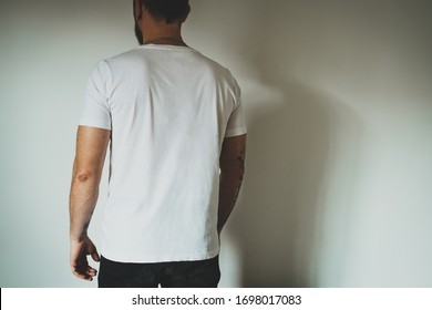 Young hipster man wearing blank white t-shirt with empty space for logo, text or design. mock-up of t-shirt, white wall in the background. Back view.