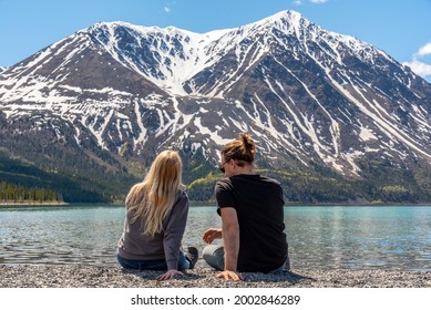 Young Hipster Man Woman Couple Sitting Lakeshore, Beside A Lake With Epic, Scenic Mountain Background In Northern Canada, Yukon Territory In Kluane National Park. 