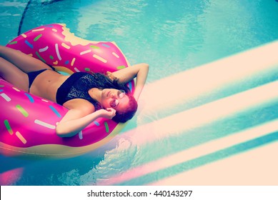 Young hipster millennial girl in sprinkled donut float at pool, festival, hotel, beach, event smiling with sunglasses on during summer