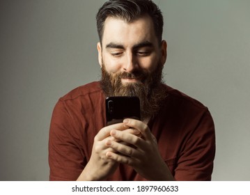 A Young Hipster Style Man With A Beard And Modern Haircut Smiling While Watching His Phone Screen With Copy Space