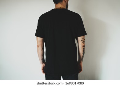 Young hipster wearing black jeans and blank black t-shirt with empty space for  logo, text or design. mock-up of t-shirt, white wall in the background. Back view.
