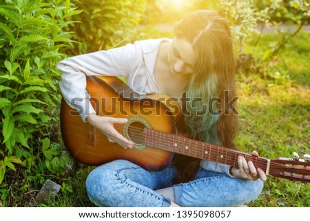 Young hipster woman sitting in grass and playing guitar on park or garden background. Teen girl learning to play song and writing music. 