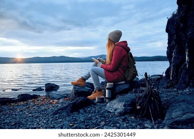 Young hipster woman sitting on the stone lake shore near the water and bonfire, wearing red coat, she is making notes in a notepad, looking at the sunset over the mountains. Solo traveler, dreamer  - Powered by Shutterstock