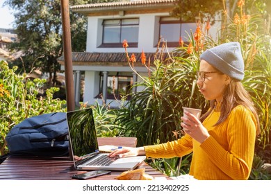 Young Hipster Woman Sitting At A Table Outside Checking Her Laptop While Drinking A Hot Drink On A Sunny Day