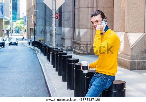 Young Hispanic American with hair bun, wearing
glasses, yellow long sleeve T shirt, blue jeans, holding cup of
coffee, sitting on street in New York City, talking on cell phone,
taking work break.