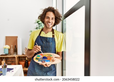 Young hispanic artist man smiling happy leaning on the window at art studio.