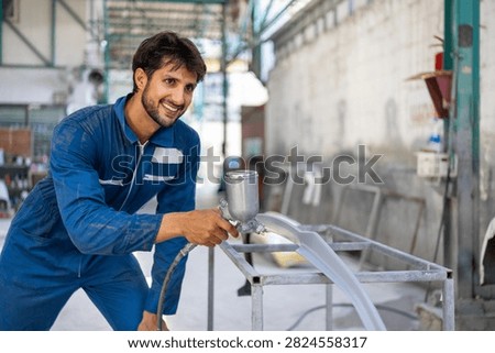A young Hispanic auto mechanic smiles at the camera while holding a spray gun near a car bumper.